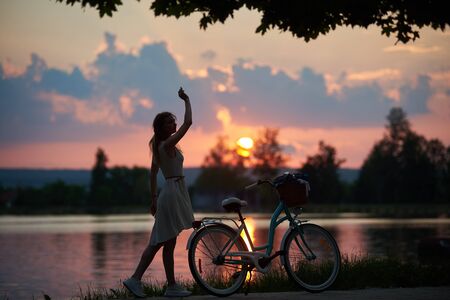Tender woman in a dress posing with a vintage bicycle near the river against the backdrop of a bewitching sunset. Warm evening in the nature. Freedom lifestyleの写真素材