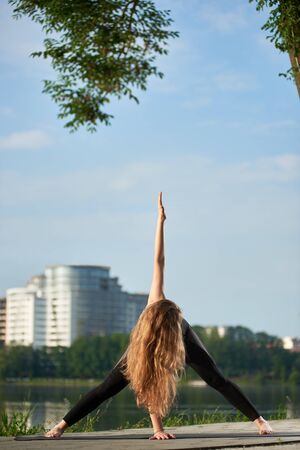 Young woman with beautiful long hair doing yoga in black sportswear under morning blue sky against city view, stretching in triangle pose, copy spaceの写真素材