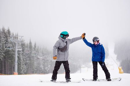 Smiling woman and man rejoicing and giving high five. Happy couple standing on snowboards on snow-covered slope with chairlift on side along coniferous forest. Dense foggy mountain views on backgroundの写真素材