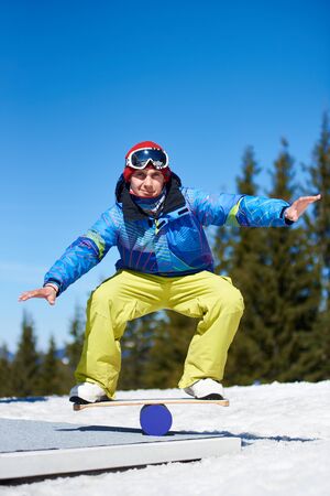 Smiling athletic male snowboarder training, balancing on snowboard on snowy hill on background of blue sky and spruce trees on sunny day. Winter extreme mountain sports, active lifestyle concept.の写真素材