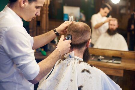 Young barber working with electric razor making haircut for stylish bearded man sitting in hairdresser chair in front of big mirror in barbershop. Back view of client. Blurred reflection in mirror.の写真素材