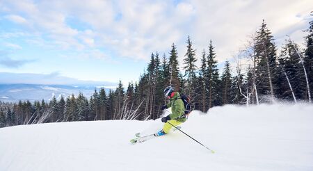 Proficient skier skiing on ski slope. Man riding on his skis very fast, making carved turns on top edge. Risk, extreme, thrill, achievement concept. Picturesque mountain view. White clouds on blue skyの写真素材
