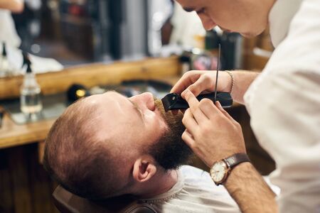 Using of electric trimmer machine for styling mustaches and beard. Accuracy and concentration of professional barber. Work process in modern hairdressing salon for men. Cropped close up view.の写真素材