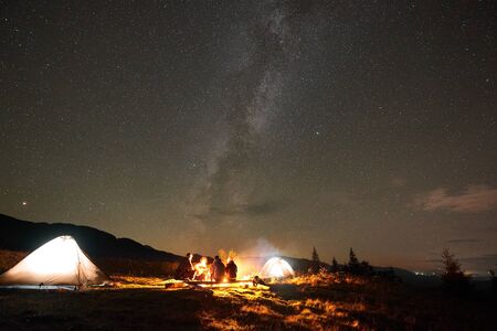 Night camping under stars. Group of four hikers, men and woman with guitar sitting by burning campfire at illuminated tourist tents under summer night starry sky and Milky Way constellation.の写真素材