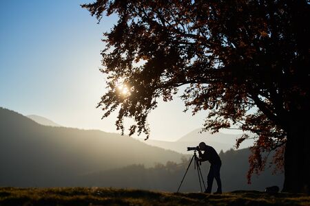 Silhouette of male tourist photographer using tripod and professional camera to take picture of mountain panorama, standing under large tree on woody foggy mountains landscape and blue sky background.の写真素材