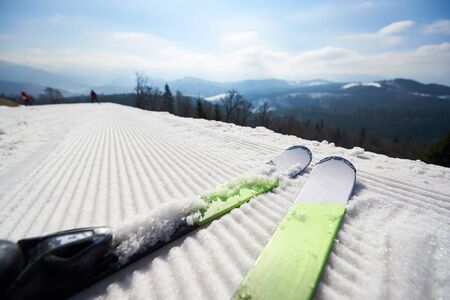 Winter landscape. Close-up of skis on track, small figures of people skiing, beautiful woody mountains with snowy peaks and blue sky background. Sports and recreation, beauty of nature concept.の写真素材