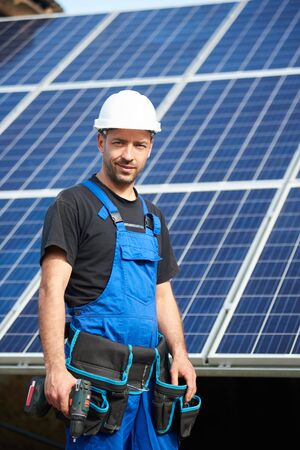 Portrait of happy engineer technician in white protective helmet and blue overalls with electrical screwdriver on background of exterior solar panel photo voltaic system blue shiny surface.の写真素材