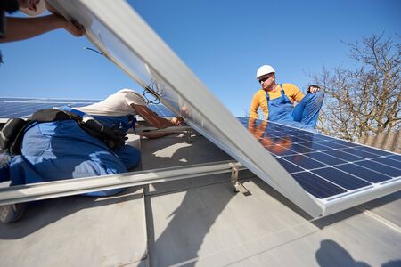 Male workers installing stand-alone solar photovoltaic panel system. Electricians mounting blue solar module on roof of modern house. Alternative energy ecological concept.の写真素材