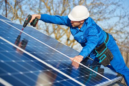 Man worker in blue suit and protective helmet installing solar photovoltaic panel system using screwdriver. Electrician mounting module on roof of modern house. Alternative energy ecological concept.の写真素材