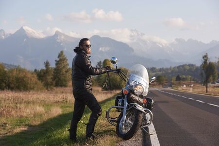 Bearded tall athletic motorcyclist in dark sunglasses, black leather clothing standing at modern powerful motorbike on background of green rural landscape, steep mountain peaks and bright sky.の写真素材