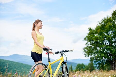 Attractive happy girl cyclist standing on a grassy hill with yellow mountain bicycle, enjoying summer day in the mountains. Outdoor sport activity, lifestyle conceptの写真素材