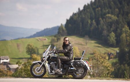 Side view of handsome bearded biker in black leather jacket and sunglasses sitting on modern motorcycle on roadside on blurred background of foggy spruce forest, green hills and bright sky.の写真素材