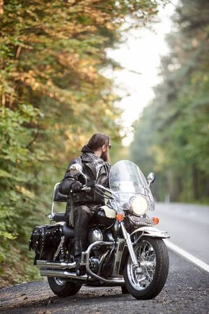 Bearded biker in black leather clothing sitting on cruiser motorcycle on country roadside on background of empty straight asphalt road and green trees bokeh foliage.の写真素材