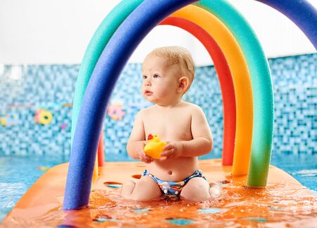 Cute playful little boy holding his rubber toy, sitting on a floating colorful construction in the swimming pool. Concept of healthy childhoodの写真素材