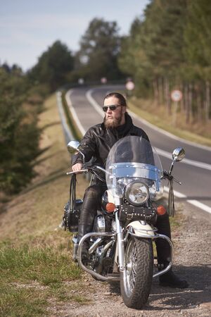 Handsome bearded biker with long hair in black leather jacket and sunglasses sitting on modern motorcycle on roadside, on blurred background of empty twisty road with white marking on sunny summer dayの写真素材