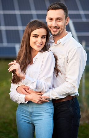 Close-up portrait of young and beautiful smiling couple, standing close to each other, holding hands, embracing, wearing white shirts, solar panel on background, concept of happy relationshipsの写真素材