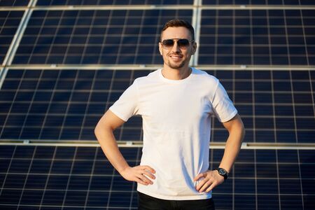 Portrait of a young successful man standing smiling with his hands on waist, wearing white t-shirt and dark sunglasses, in front of a big solar pv moduleの写真素材