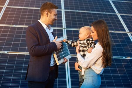 Cheerful kid in his mother arms examines the solar battery that dad holds, against background solar panels. Concept of family warmth and comfort in modern world with continuously advancing technologyの写真素材