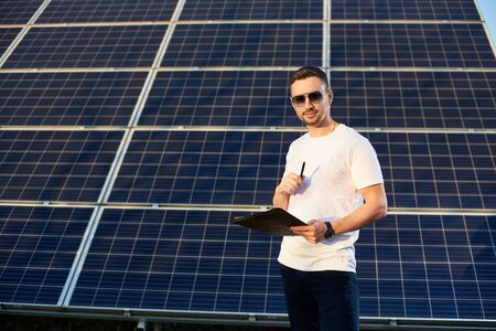 Handsome male with a folder and pen in his hands on a background of solar panels on a warm day. Young man in sunglasses, white T-shirt and jeans is looking at the camera with a smileの写真素材