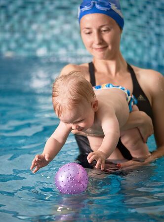 Close-up snapshot of mother holding her baby boy in her arms in indoor pool. Blonde kid is trying to reach for his purple inflatable ballの写真素材