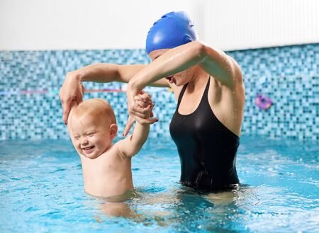Young mother wearing a cap and swimming suit is doing exercises with her cheerful little son in the swimming pool holding his both hands up. Concept of healthy family leisureの写真素材