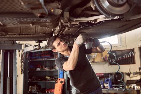 Repairman fixing car in garage. Experienced specialist car mechanic standing under lifted car during repair and maintenance process in repair station. Workshop for automobile checking up and repairingの写真素材