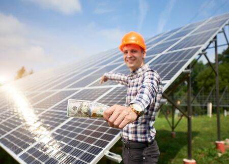 Close up hands mans with hundred dollar bill in it and pointing at the background of solar station. Business and generation concept. Green ecological power energy generation.の写真素材