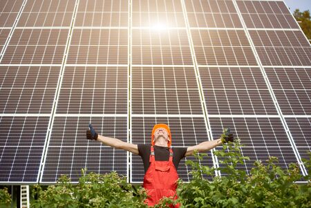 Smiling worker with solar station, raising his hands, showing thumbs up on a background of photovoltaic panels near the house. Man in orange uniform. Science solar energy. Renewable energy.の写真素材