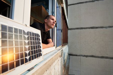 Young man on balcony of residential building with solar panel in which the sun is reflected at sunset. Concept converting solar radiation into electricityの写真素材