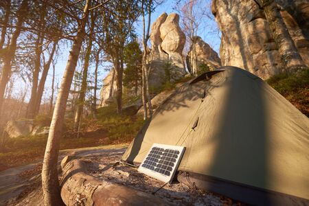 Small solar panel and tourist tent in the forest at the foot of the cliffs. Fir trees and fallen leaves on a sunny day surround the tent camp which is heated by a solar batteryの写真素材