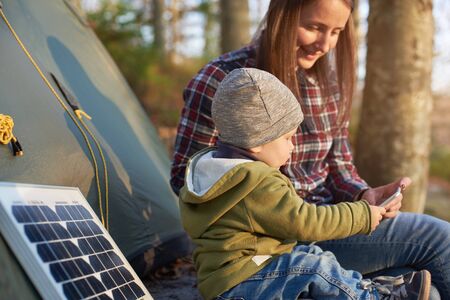 Little wonderful kid takes the phone from a mother sitting near the solar panel at a campsite in the forest under the suns rays. Child dressed in jeans, jacket and hat in the middle of warm autumnの写真素材