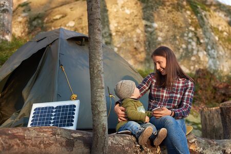 Mother smiling to child, sitting on a log in the forest near tourist solar panel and a tent camp. Woman is holding a telephone. Family weekend in the warm autumn evening outdoorの写真素材