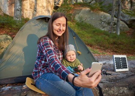 Woman and a child take a selfie on a mobile phone on the background of a tent with a solar battery at the foot of a cliff in the forest. Family in the sunbeams with gentle smiles looking at the cameraの写真素材