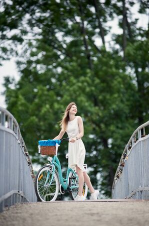 Young girl in a white dress is standing on the bridge with her blue citybike, summertime, smiling face to the wind, green trees on backgroundの写真素材