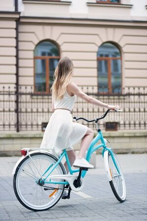 Nice girl in light white dress is riding a blue bike on the street in the city looking up at the building, pavement, full length, back viewの写真素材