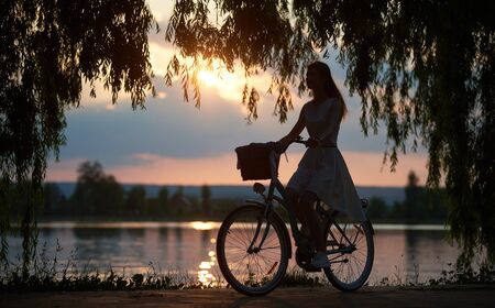 Silhouette of a girl riding a bike near urban lake at beautiful summer sunset, willow branches on the foreground,romantic sky on the background, copy spaceの写真素材
