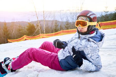 Close-up snapshot of a girl lying on the snow at ski resort, looking at camera, wearing ski outfit, helmet, glasses, gloves and snowboard, side viewの写真素材