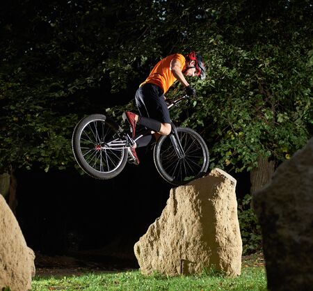 Back view of cyclist in sportswear jumping on big rock. Sporty man riding a mountain bike at night in city. Concept of night workout.の写真素材