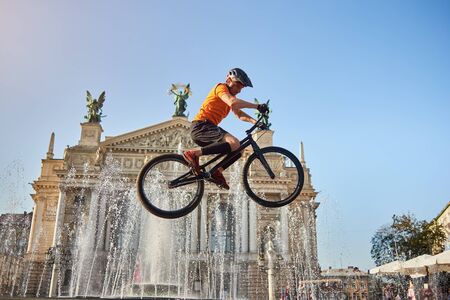 View from below active guy in orange shirt holding black bicycle on architecture background. Extreme athlete in black helmet performing a stunt in front of a fountain. Concept of stunt in air.の写真素材
