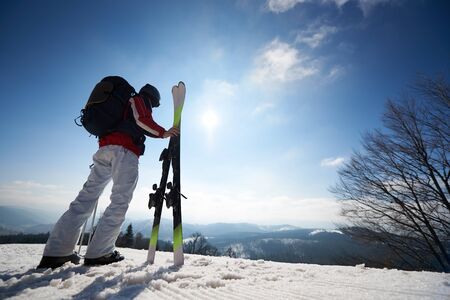 Back view of man skier with skis, poles and backpack standing in white snow, beautiful winter mountain landscape and blue sky copy space background. Sports and recreation, active lifestyle concept.の写真素材