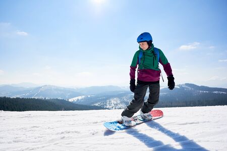 Young female snowboarder riding snowboard on copy space background of clear bright blue sky and snowy mountains on sunny winter day. Winter sports and recreation, leisure outdoor activities concept.の写真素材