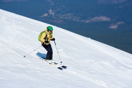 Professional skier in helmet with backpack riding skis fast down steep snowy mountain slope on background of deep white snow and green spruce trees. Winter holidays, sport and recreation concept.の写真素材