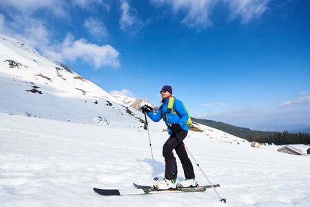 Handsome tourist in colorful clothing and sunglasses with backpack trekking on skis in deep snow on background of bright blue sky and beautiful mountain. Winter vacations, active lifestyle concept.の写真素材