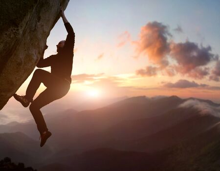 Rock climbing. Young male risky climber trying staying on challenging cliff route. Scenic mountain landscape with sunrise sky on background. Fail, difficulties, overcoming obstacles. Copy spaceの写真素材
