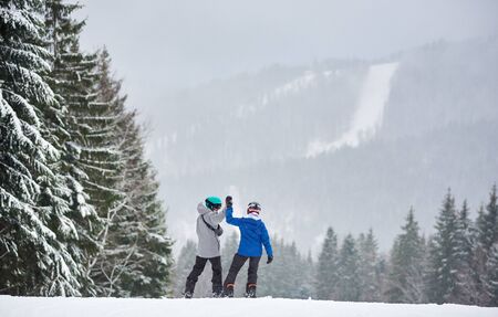 Couple of snowboarders with hands up preparing descending from high slope with charming forest around during snowing in mountains. Back view. Happiness, achievement, support, friendship conceptの写真素材