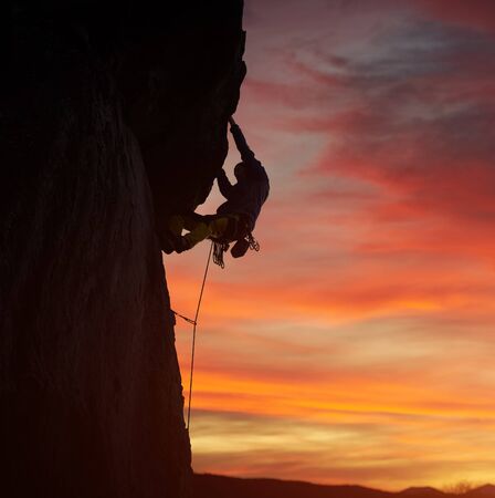 Male silhouette of proficient persistent climber in safety harness climbing at nightfall and pulling up on rock with both hands. Copy space on sunset orange cloudy sky. Snapshot with low angle viewの写真素材