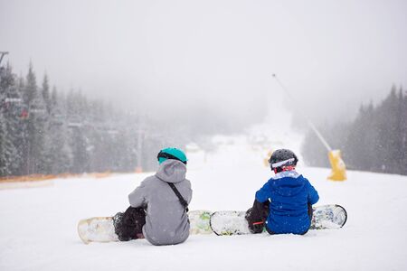 Back view of pair of snowboarders with boards on sitting on snow-covered slope in winter snowfalling weather. Couple looking at each other on wooded mountain hill. Fear, uncertainty, support conceptの写真素材