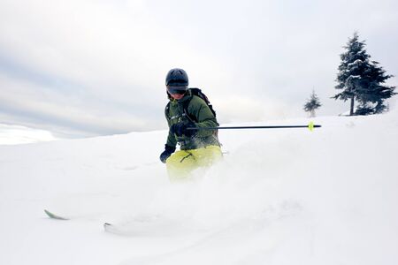 Skier male into deep white snow powder on snow-covered high mountain peak. Extreme skiing in winter sports season. Dangerous, activity, thrill concept. Grey sky and pine tree on background. Front viewの写真素材