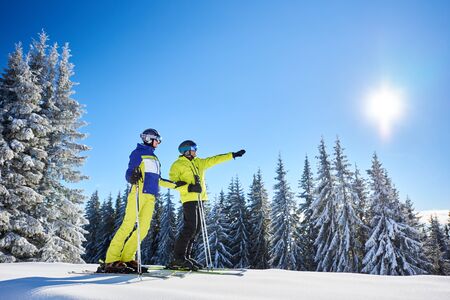 Low angle shot of skiers resting during skiing in sunny winter weather. Male skier showing sun high in blue sky over winter mountains. Fantastic recreational area of ski resort on background.の写真素材