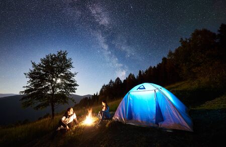 Happy romantic couple tourists having a rest at bonfire beside camp and blue glowing tent under amazing night sky full of stars and Milky way. On background magical starry sky, mountains and big treeの写真素材